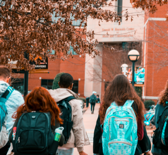 A group of students with backpacks walk on a college campus pathway lined with autumn trees and a brick building in the background.