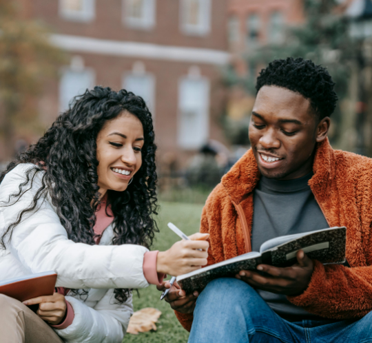 Two people sitting outdoors on grass, smiling and looking at a notebook together while one writes and the other holds the notebook.