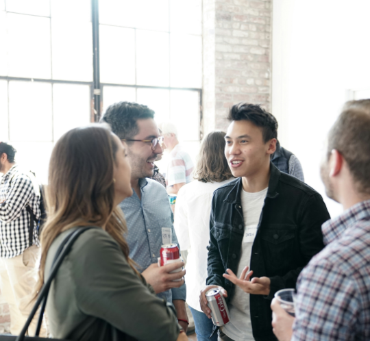 A group of four people stand indoors, holding drinks and talking near large windows with natural light.